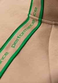 Tissu beige avec une bande bordée de vert comportant le mot « performance » brodé plusieurs fois le long de la couture.