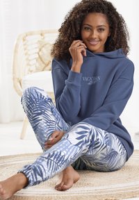 Felpa con cappuccio blu con stampa "VACANZA"; pantaloni jogger blu e bianchi a fantasia con foglie di palma; modello seduto su un tappeto rotondo testurizzato.
