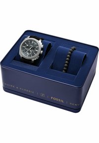 Fossil-ur og sort lavastensarmbånd i en marineblå æske med guldfarvet skrift. Uret har en sølvfarvet kasse og en tekstureret mørk skive.