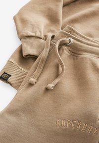 Beige sweatshirt och byxor set, med ribbade muddar och dragsko. Huvan har en texturerad logotyp på framsidan. Slätt tyg.