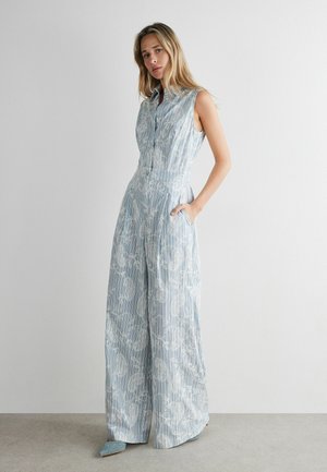 Vrouw draagt een mouwloze blauw-wit gestreepte jumpsuit met bloemenpatroon, staand met één hand in de zak tegen een effen muur.