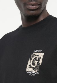 Camiseta de algodón negra con un destacado gráfico beige que presenta una "G" estilizada en medio de intrincados patrones florales y de paisley.