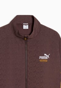 Sötétbarna Puma King dzseki geometrikus mintával, magas gallérral és teljes cipzárral. A mellrészen fehér logó sárga részletekkel.