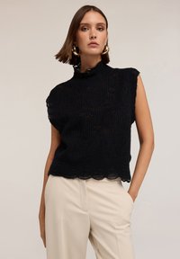 Top negru fără mâneci din tricot, cu guler înalt, model texturat și margine fantezistă. Purtați-l cu pantaloni de culoare deschisă, evidențiind o croială lejeră.