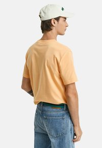 Kurzärmliges, pastellorangefarbenes T-Shirt aus Baumwolle, kombiniert mit hellblauen Jeans und einer weißen Kappe mit grünem Text. Rückansicht.