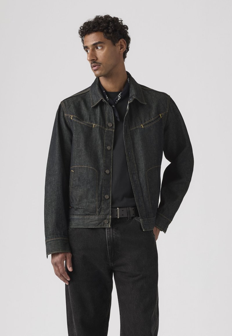 Mand med krøllet hår iført en sort denimjakke, sorte bukser og sort bælte, der ser mod venstre mod en ensfarvet baggrund.