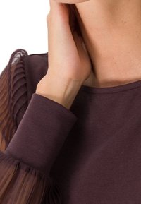 Mørk lilla pullover med ribbet struktur og transparente puffærmer. Nærbillede af halsudskæringen og håndleddet, der viser det glatte stof.