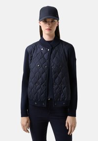 Granatowa pikowana kurtka bombka z zatrzaskami, prążkowanym kołnierzem i rękawami; noszona z dopasowaną granatową czapką. Gładka tekstura, dopasowany krój.