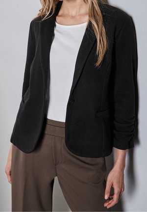 Žena v černém jednoprávkovém saku přes bílý top a hnědé kalhoty stojí před neutrálním šedým pozadím.