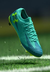 Τυρκουάζ ποδοσφαιρικό παπούτσι με υφή στην επάνω επιφάνεια, δυναμικές ρίγες και κίτρινες λεπτομέρειες. Διαθέτει σύστημα δέσιμου με κορδόνια και εξωτερική σόλα με τάπες για καλύτερη πρόσφυση.