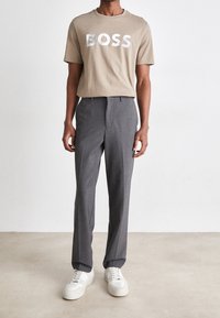 Muž v béžovém tričku s nápisem "BOSS", šedých kalhotách na míru a bílých teniskách, stojící na jednobarevném bílém pozadí.