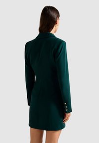 Donna con lunghi capelli castani che indossa un abito blazer verde scuro con bottoni dorati sui polsini, vista di spalle contro uno sfondo grigio.
