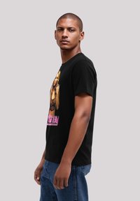 Schwarzes Baumwoll-T-Shirt mit einem grafischen Druck einer Figur und pinkem Text. Kurze Ärmel, Rundhalsausschnitt, lockere Passform. Sichtbare Jeans aus Denim.