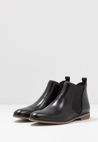 Botas de tobillo de cuero negro con paneles laterales elásticos y tiradores. Forma delgada, punta redondeada y un tacón de madera bajo. Textura suave.