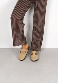Zapatos de ante sin cordones en beige con detalles troquelados y una hebilla decorativa, combinados con pantalones sueltos de rayas marrón y blanca.