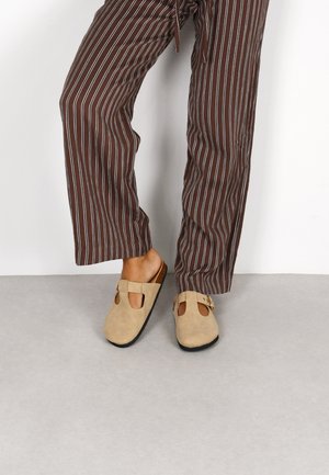 Zapatos de ante sin cordones en beige con detalles troquelados y una hebilla decorativa, combinados con pantalones sueltos de rayas marrón y blanca.