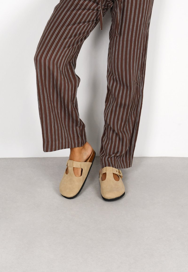 Zapatos de ante sin cordones en beige con detalles troquelados y una hebilla decorativa, combinados con pantalones sueltos de rayas marrón y blanca.
