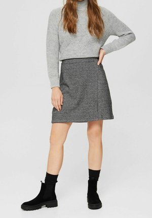 Femme portant une jupe grise avec motifs, un pull en maille gris, des chaussettes noires aux chevilles et des bottines en daim noires, debout avec une main sur la hanche.