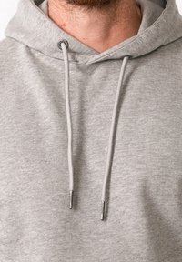 Sudadera gris hecha de tela suave con capucha ajustable por cordón. Presenta ojales de metal y puntas de cordón en contraste. Diseño simple con textura mínima.