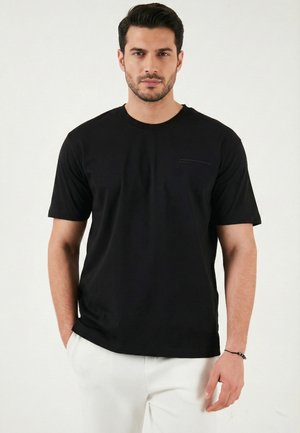 Homme aux cheveux foncés et à la barbe portant un t-shirt noir uni à manches courtes et un pantalon blanc, debout devant un fond blanc, les mains le long du corps.