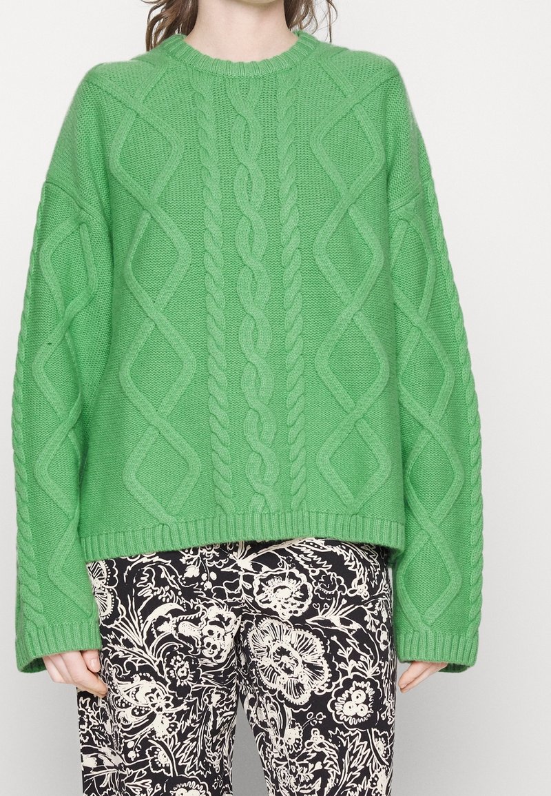 Pull vert en maille avec motifs torsadés et losanges ; col rond et coupe oversize. Associé à un pantalon imprimé floral noir et blanc.