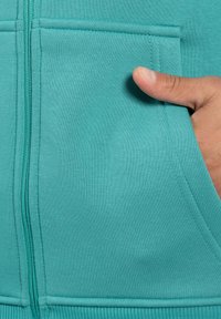 Felpa con zip color teal, con una mano parzialmente all'interno della tasca frontale. Presenta polsini a costine e una texture liscia con dettagli di cucitura tono su tono.