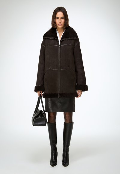 Manteau en shearling noir avec un large col, fermeture éclair sur le devant et finition texturée. Associé à une jupe en cuir noire et des bottes montantes jusqu'aux genoux.