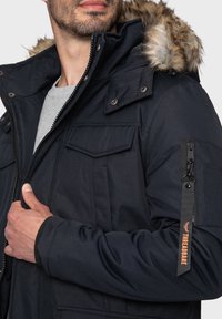Námořnická parka s kapucí s kožešinovým podšívkou, která má mnoho kapes, zipové zapínání a elastické manžety. Mezi kovovými prvky jsou patentky a zipy.