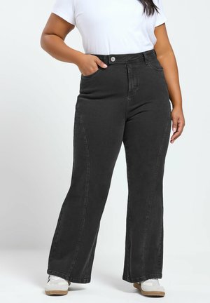 Frau trägt hoch taillierte schwarze Jeans mit weitem Bein, weißes T-Shirt und weiße Sneaker, steht mit einer Hand in der Tasche.