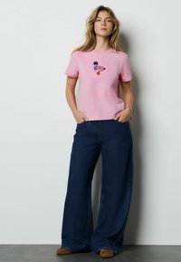 Roza bombažna majica s pisanim vezenim vzorcem metulja, kombinirana s širokimi temno modrimi kavbojkami in rjavimi obuvali brez vezalk.