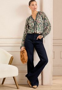 Srajca s cvetličnim vzorcem in prosojnimi rokavi, v kombinaciji z visokim pasom temno modrimi flare hlačami; drži tan sueden clutch.