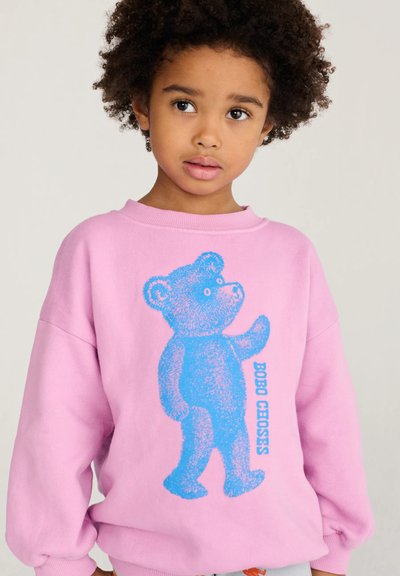 Enfant aux cheveux bouclés portant un sweat-shirt rose avec un grand dessin d'ours en peluche bleu et le texte "BOBO CHOSES" sur le devant.