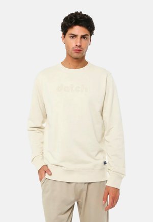 Uomo che indossa una felpa beige e pantaloni kaki chiari, in piedi con una mano in tasca davanti a uno sfondo bianco uniforme.