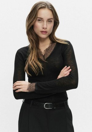 Svart långärmad topp med en spetsdetalj vid V-ringningen och ärmsluten, tillverkad av ett genomskinligt, lätt tyg. Matchas med ett svart bälte.