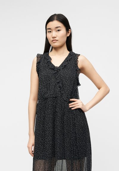 Νεαρή γυναίκα με ίσια σκούρα μαλλιά που φορά ένα μαύρο αμάνικο φόρεμα με λευκά πουά και βολάν στο ντεκολτέ, χέρι στη μέση.