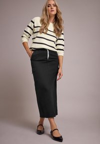 Gonna maxi in denim nero con tasca laterale, abbinata a un maglione crema con righe nere e scollatura a V. Scarpe nere flat.