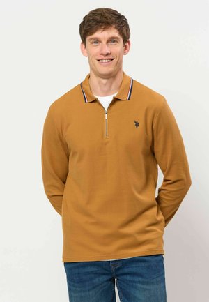 Hombre sonriente con un suéter de cuello con cremallera color mostaza y vaqueros azules, de pie frente a un fondo claro y liso.