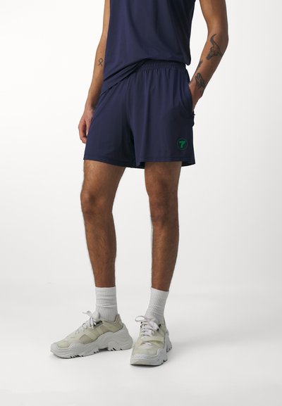 Marine idrettsshorts med elastisk midjebånd, sidelommer og en grønn "7"-logo på venstre lår. Brukt med lyse sneakers og sokker.