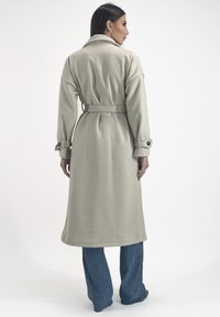 Trench coat beige chiaro con una finitura testurizzata, caratterizzato da un ampio colletto, vita con cintura e polsini con bottone. Indossato sopra jeans blu a gamba larga.