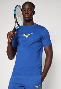 Zils īsām piedurknēm sporta krekls ar dzeltenu Mizuno logo uz krūtīm, ko valkā persona, kas tur tenisam raketi. Gluda auduma tekstūra.