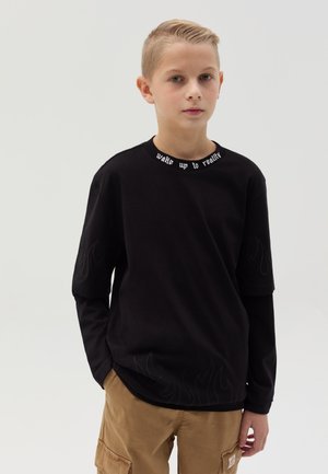 Zwarte T-shirt met korte mouwen met een vlammenpatroon aan de onderrand en een witte slogan op de kraag, gecombineerd met khaki cargobroeken.