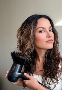 Sèche-cheveux avec un design noir ergonomique et un large embout, tenu par une personne ayant des cheveux ondulés descendant aux épaules. Texture lisse et finition brillante.