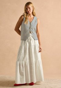 Bezrukávový pruhovaný top v námorníckej modrej a bielej farbe s gombíkmi; viacvrstvová biela maxi sukňa s očkovými vzormi a volánkovým lemom; červená obuv.