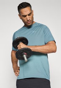 Camiseta deportiva en color azul verdoso con rayas horizontales, mangas cortas, escote redondo y un diseño ajustado, acompañada de una mancuerna negra.