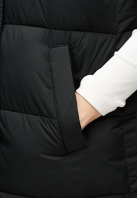 Mano en el bolsillo de una chaqueta acolchada negra, con una camiseta de manga larga blanca con ribetes.