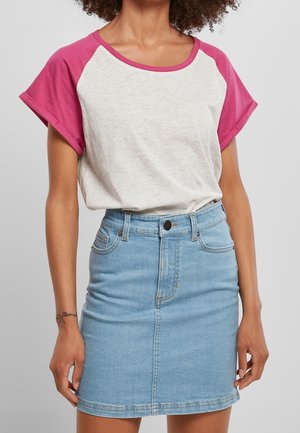 Vrouw die een wit- en roze shirt met korte mouwen draagt, gestopt in een lichtblauwe high-waist spijkerrok, staand tegen een effen achtergrond.