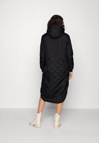 Abrigo largo negro acolchado con capucha, con un patrón diagonal y aberturas laterales, combinado con botas beige de cordones.