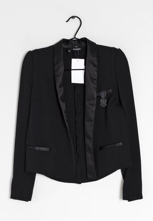 Blazer noir sur mesure avec revers en satin, deux poches avant, une épingle décorative sur la poche poitrine, et une étiquette blanche, suspendu à un cintre noir.