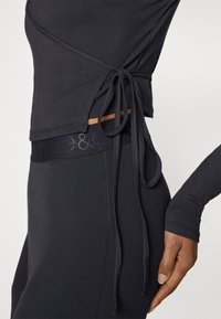 Fekete bordázott felső hosszú ujjakkal, oldalt megkötővel. Magasderekú fekete leggingsszel párosítva, amelyen finom logószalag látható.