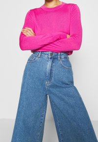 Haut en maille fuchsia à manches longues associé à un jean denim bleu taille haute à jambes larges, avec poches avant et fermeture classique à boutons.